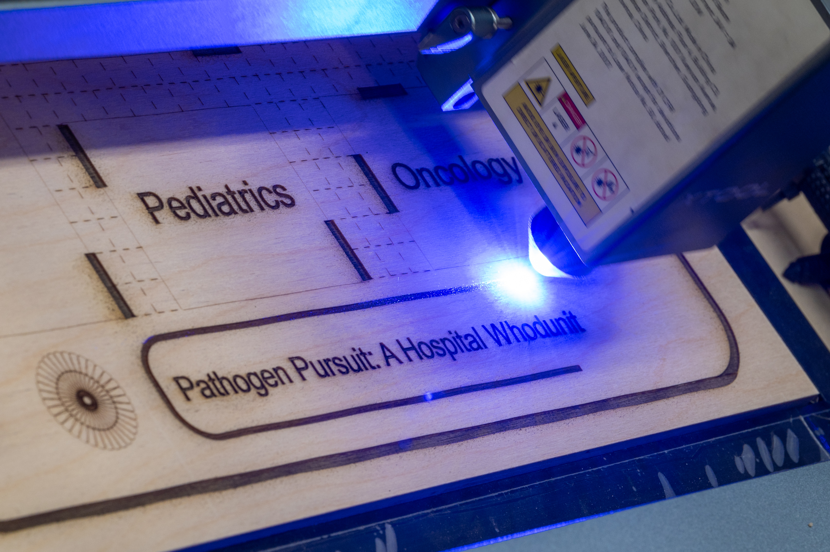 A laser cutting a piece of plywood that shows "Pathogen Pursuit: A Hospital Whodunit"